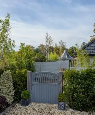 En gråmalet træport omgivet af hække, med grusgulv.