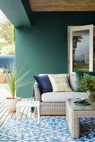 Design af en gårdhave i en blågrøn indretning med terrassebord, blåmønstret tæppe og flet sofa og sofabord.
