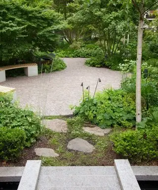 Et rundt terrassedesign i mursten i en skovklædt have med trædesten og en stenbro.