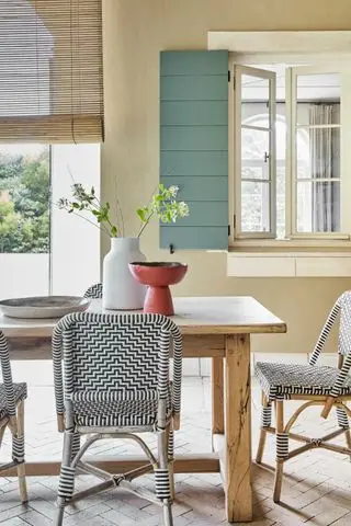 Ein hölzerner Terrassentisch und geflochtene, einfarbige Stühle vor einem cremefarbenen Haus mit Fensterläden in Eau de Nil.
