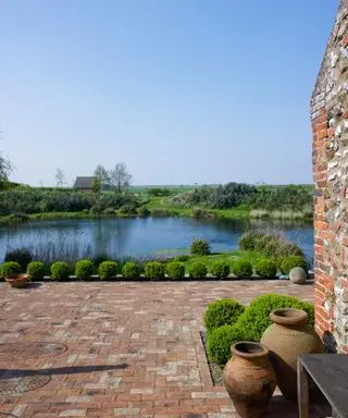 Ein mit Ziegeln gepflasterter Innenhof mit kleinen runden Hecken mit Blick auf einen See und Felder.