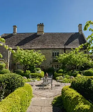 Ein formeller Innenhofgarten mit Steinpflastersteinen, Buchsbaumhecken, weißen Blumen und einem eisernen Tisch und Stuhl vor einem steinernen Landhaus.