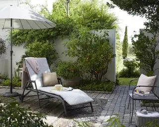 Come progettare un piccolo giardino illustrato con un lettino e un ombrellone in un giardino pavimentato, con un muro bianco e piante rampicanti.