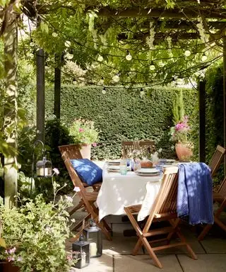 Tavolo e sedie in legno in un piccolo giardino, circondato da siepi e sotto un pergolato con glicine e fili luminosi.