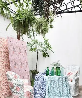 Idee per un giardino sul balcone con diversi tessuti pastello e floreali drappeggiati su un tavolo e sedie, con un albero in vaso e pareti bianche sullo sfondo.
