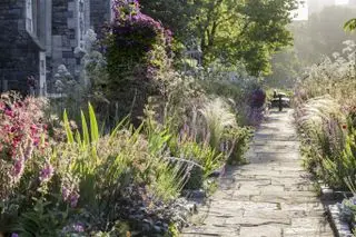 Un camino de piedra junto a un gran parterre lleno de flores violetas y plantas verdes