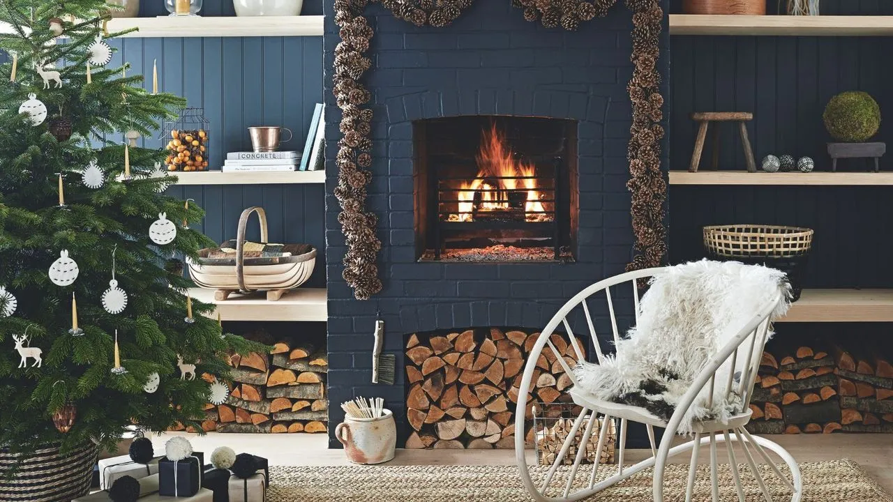 En mørkeblå stue med panelte vegger hyller og en murstein peis med tent bål. Rundt bålet er det en konglekrans og til venstre i forgrunnen er det et stort juletre dekorert med hvite ornamenter. Til høyre for den er en stor hvit statement-stol i metall med hvit, myk pute.' loading='lazy' title=