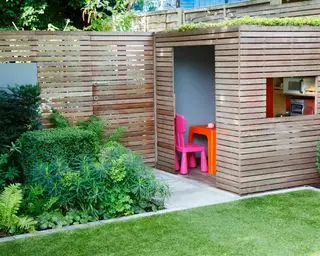 Una valla de listones de madera y un jardín detrás de un césped limpio y un parterre cuadrado de flores en un jardín urbano.