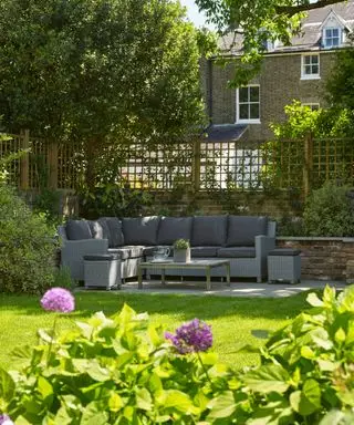 Ideas para paredes de jardín con un enrejado encima de una pared baja de ladrillos en un jardín urbano con un sofá de mimbre en esquina en forma de L, un césped soleado y aliums de color púrpura.