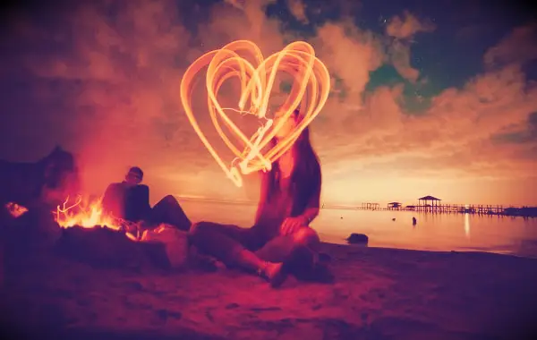 A woman making a heart in the air with a glow stick during an evening at the beach.