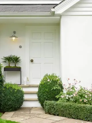 Casa con exterior en blanco y puerta de entrada blanca, camino a puerta y follaje de plantas y flores.