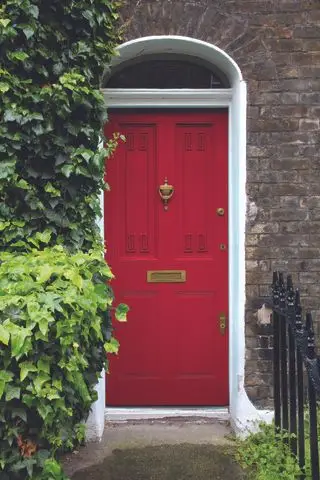 Puerta delantera roja en una casa de ladrillo con barandillas negras a un lado del camino y follaje al otro