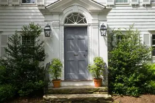 Casa con revestimiento blanco y puerta frontal azul gris con plantas de follaje a ambos lados