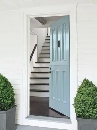 Puerta frontal de color azul claro en casa con revestimiento blanco abierta al vestíbulo y a la escalera