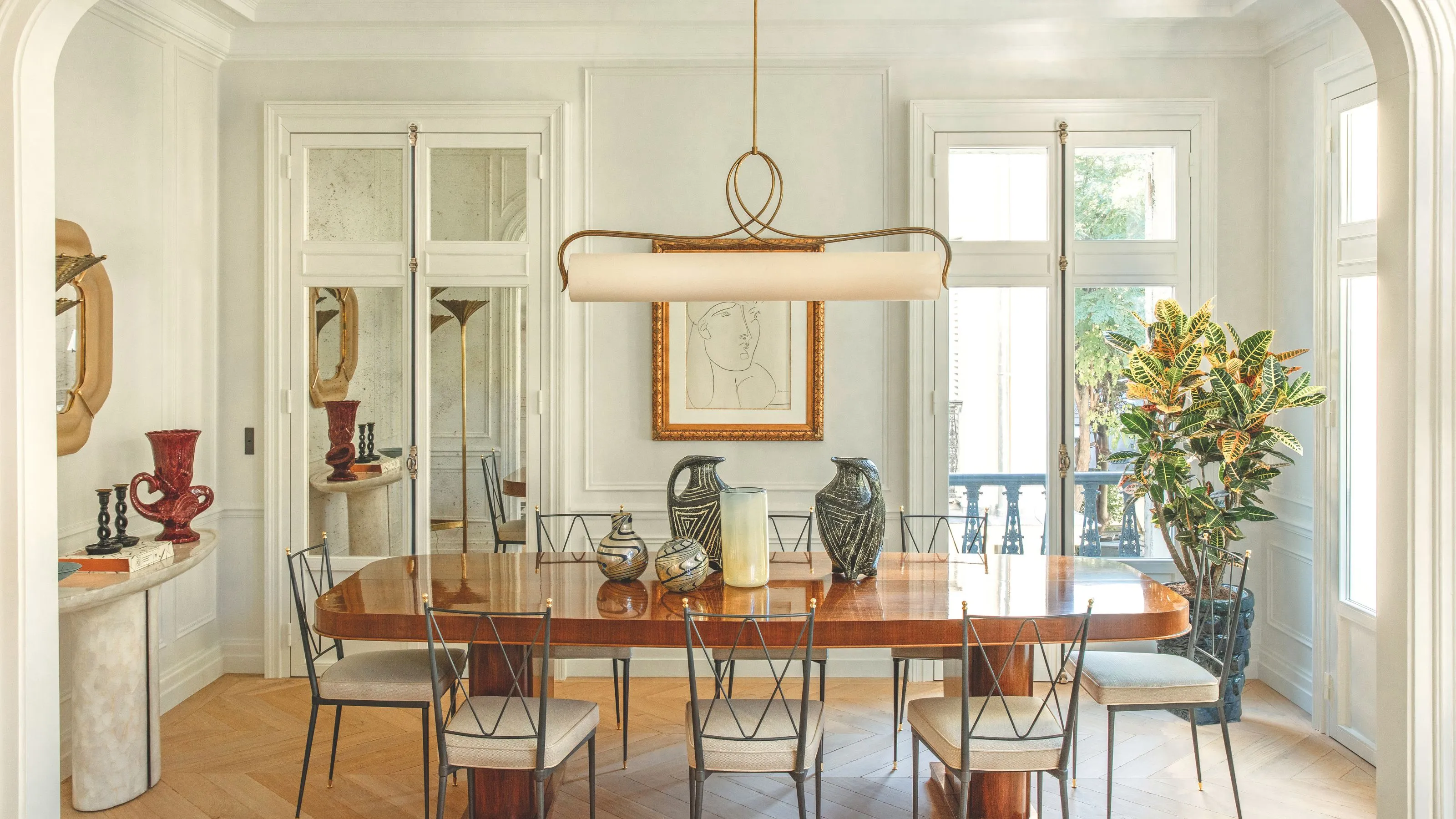 En elegant solbelyst spisestue med et bord i poleret træ, slanke sorte metalstole og en unik vandret pendel. Rummet er indrettet med keramikvaser og en stor indrammet skitse på en panelvæg.' loading='lazy' title=