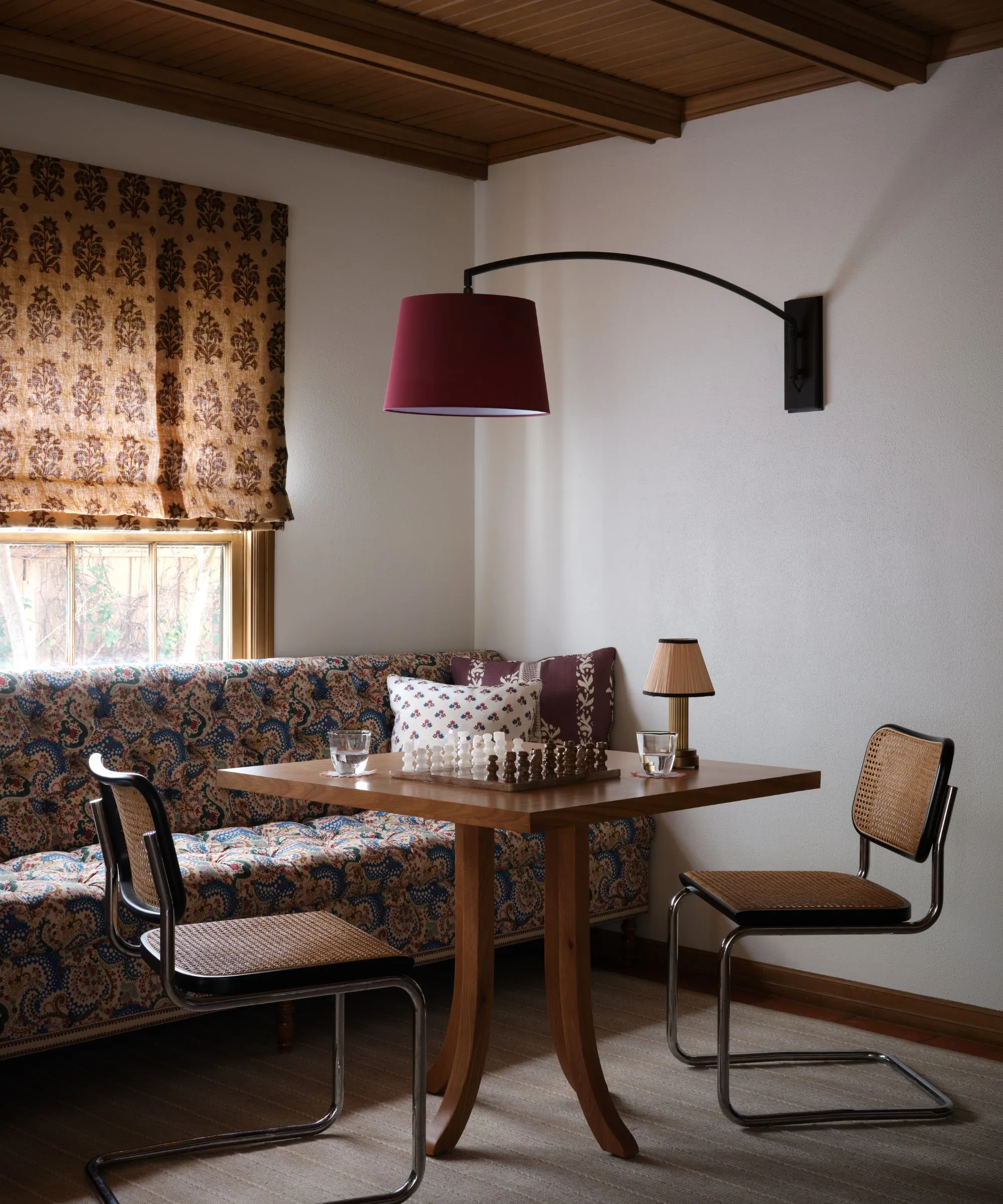 Um canto aconchegante com uma banqueta estampada, uma mesa de pedestal de madeira com um tabuleiro de xadrez e uma moderna luminária de parede com braço oscilante cor de vinho' loading='lazy' title=