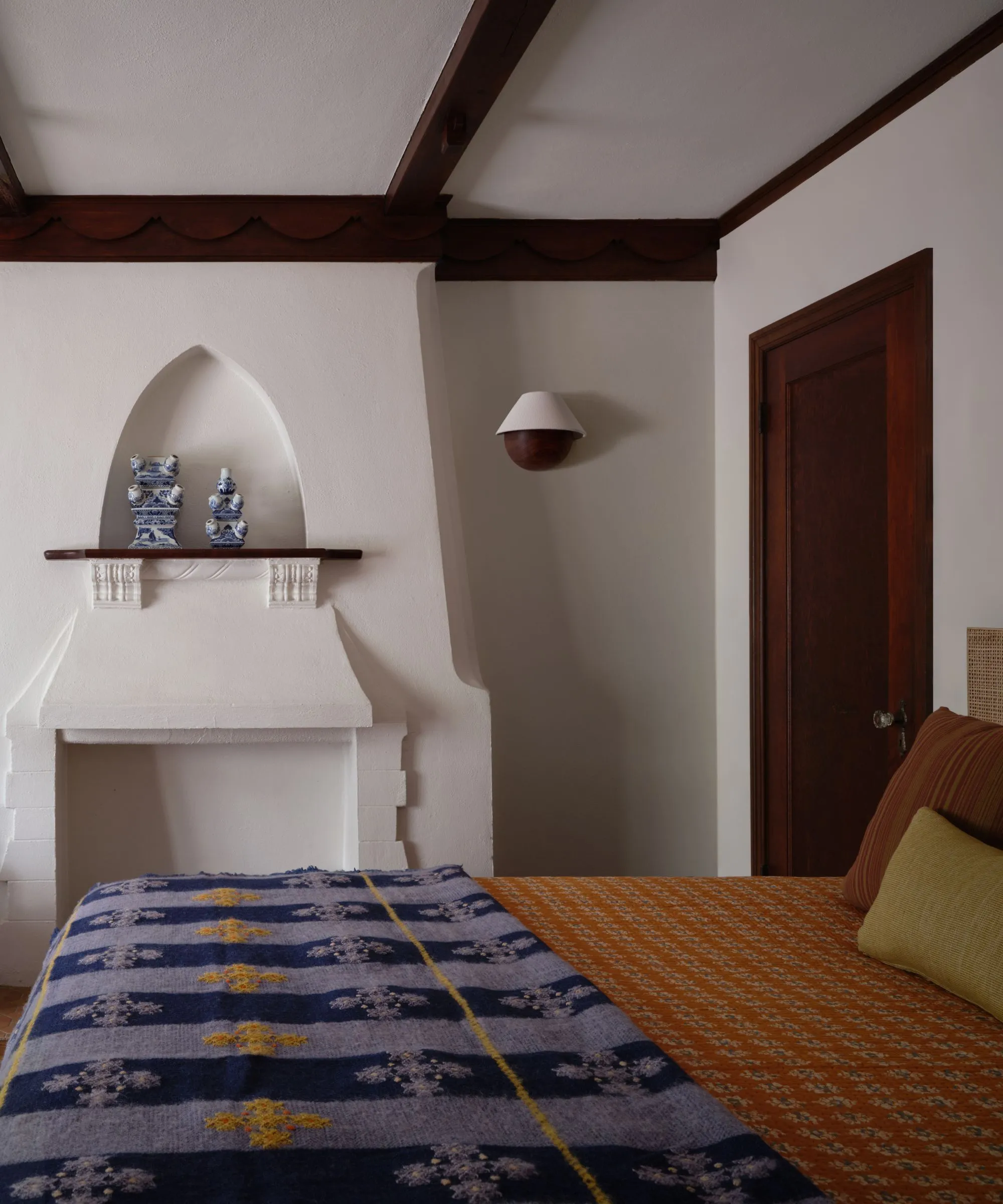 Vista do quarto mostrando uma lareira arquitetônica branca com um nicho pontiagudo acima de um cobertor estampado azul na cama e acabamento em madeira escura no teto' loading='lazy' title=