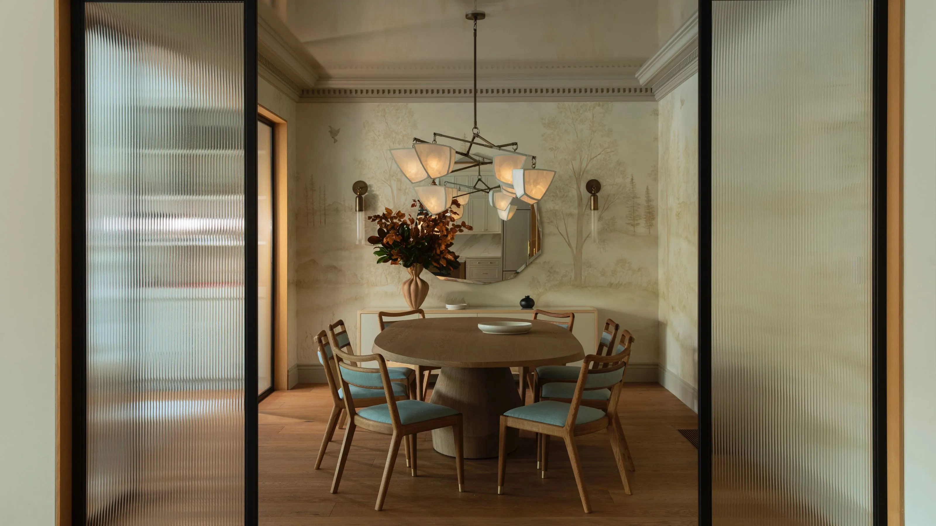 Une élégante salle à manger vue à travers des portes coulissantes en verre cannelé et dotée d'un guéridon rond en bois entouré de chaises modernes du milieu du siècle avec un revêtement bleu clair. La pièce est décorée d'un papier peint mural de paysage pittoresque, d'un grand lustre sculptural aux nuances blanches et d'un buffet minimaliste sous un miroir rond.' loading='lazy' title=