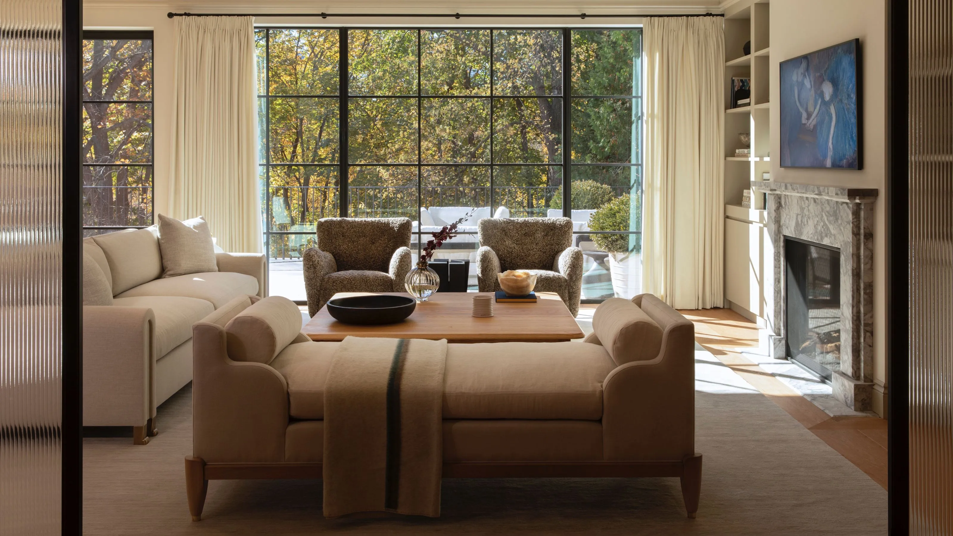 Un salon lumineux et spacieux vu à travers des portes en verre cannelé doté d'une grande fenêtre à cadre noir qui donne sur les arbres luxuriants d'automne. L'espace est ancré par un canapé neutre et deux fauteuils anthracite texturés centrés autour d'une table basse en bois avec une cheminée entourée de marbre et des étagères intégrées à droite.' loading='lazy' title=