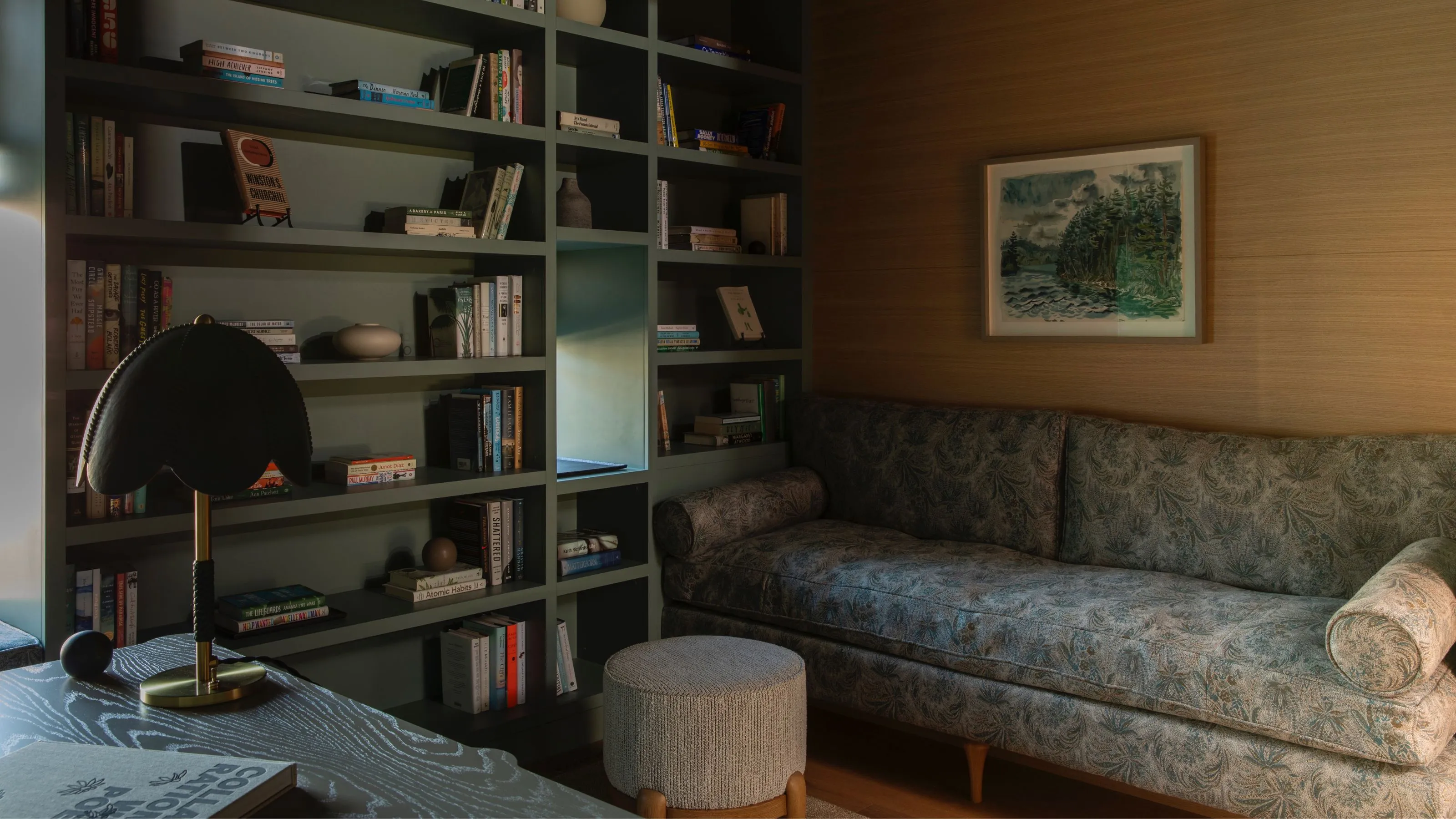 Un coin bibliothèque confortable et sombre doté d'une grande étagère intégrée vert olive remplie de livres et de petites sculptures. Un canapé moelleux à motifs floraux discrets est posé contre un mur lambrissé, accompagné d'un petit pouf texturé et d'une lampe de table noire moderne.' loading='lazy' title=
