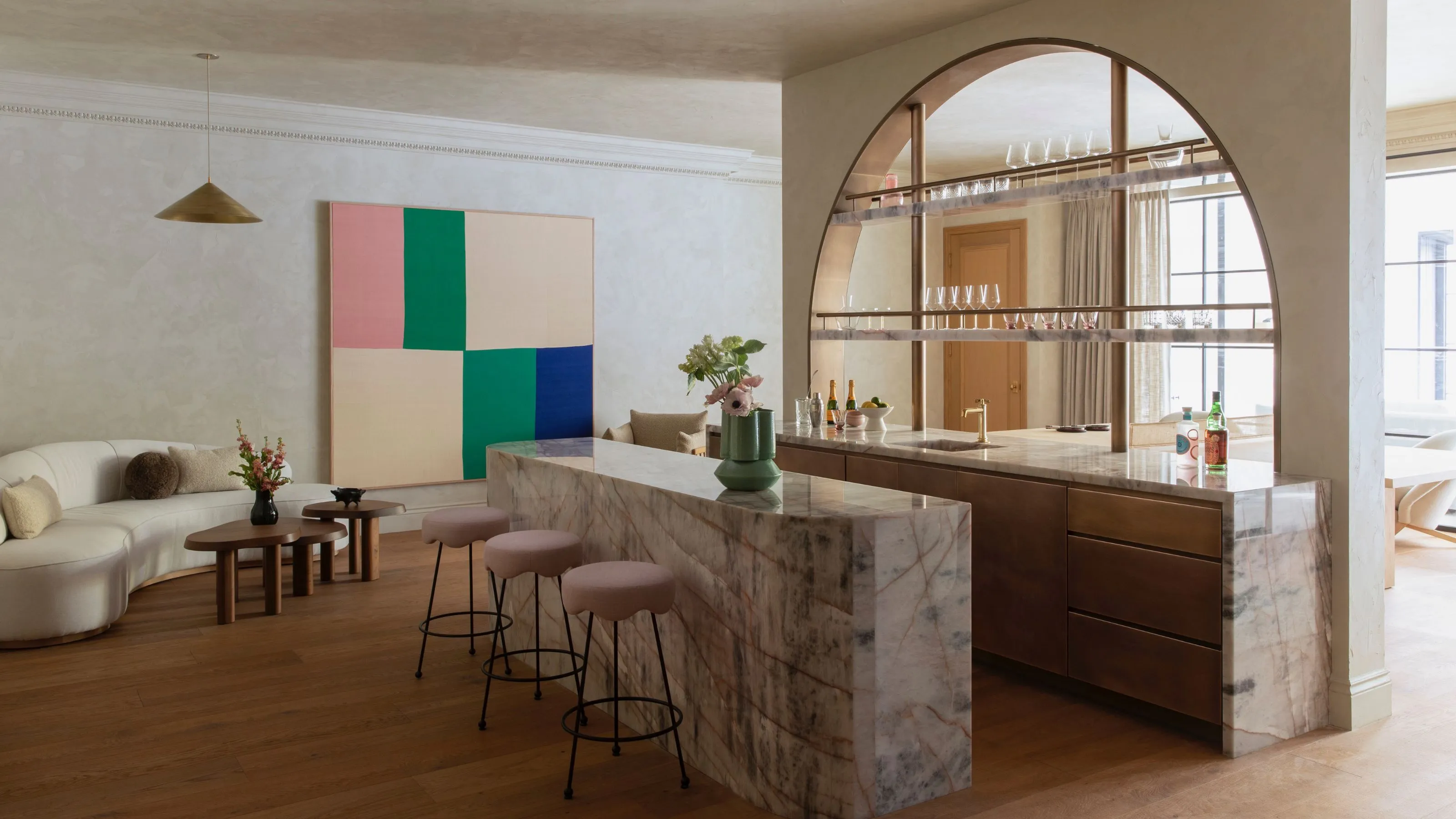 Un luxueux coin salon à aire ouverte comprenant un spectaculaire îlot en cascade de marbre veiné de violet et des tabourets de bar festonnés roses. L'arrière-plan comprend une grande ouverture architecturale cintrée avec des étagères en verre, un canapé crème incurvé et des œuvres d'art murales abstraites et vibrantes.' loading='lazy' title=