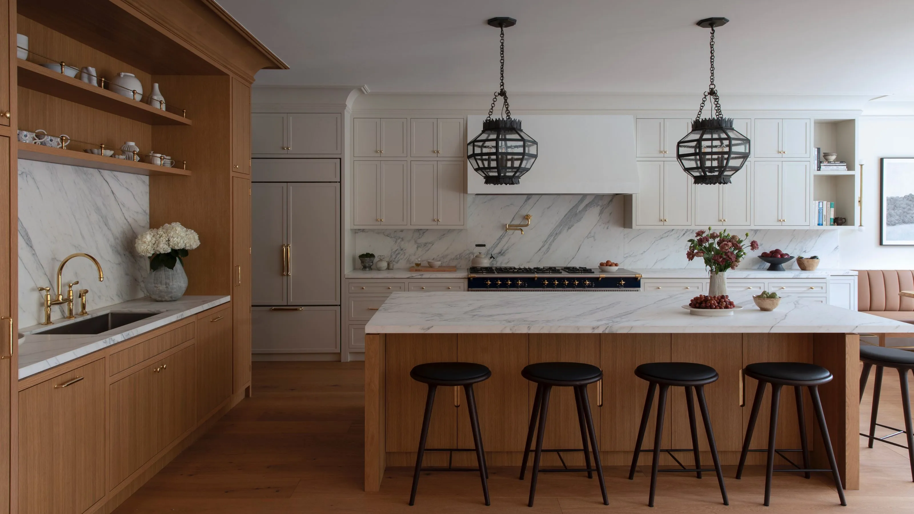 Une cuisine spacieuse haut de gamme dotée d'un grand îlot en chêne avec un comptoir en marbre blanc et des tabourets de bar minimalistes noirs. L'arrière-plan montre des armoires de couleur crème, un dosseret en marbre assorti et deux suspensions ornées de style lanterne noire suspendues au plafond.' loading='lazy' title=