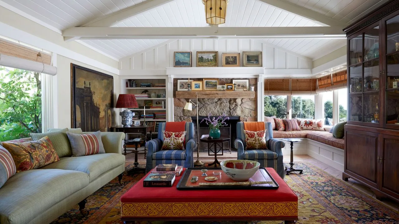 Salón con techos con vigas blancas, gran alfombra persa, otomana roja, sofá azul pálido con coloridos cojines, sillones a rayas azules y chimenea de piedra' loading='lazy' title=