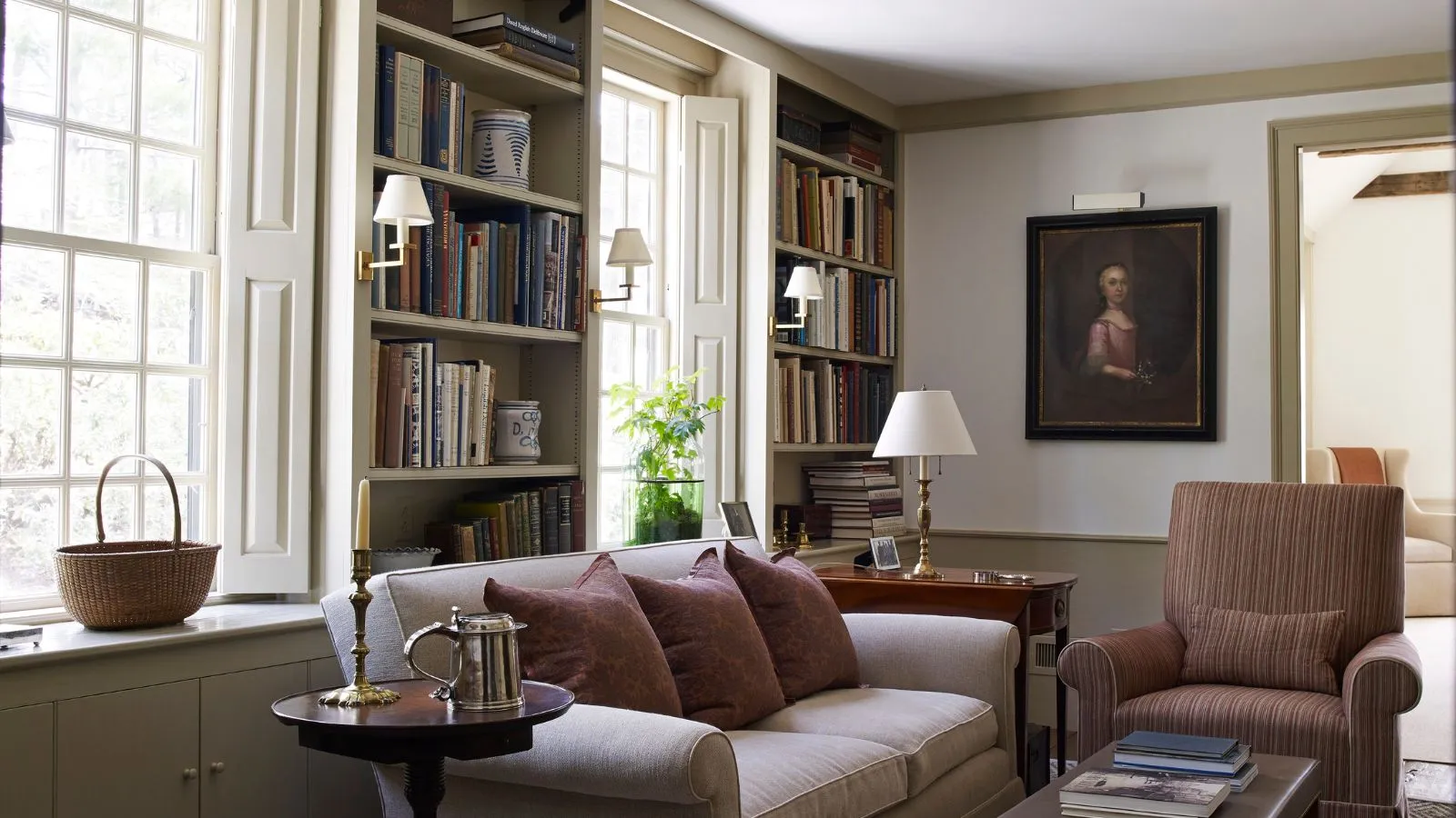 Traditionelles Wohnzimmer mit Wandlampen, großen Fenstern und Sofa mit Bücherregalen und antiken Gemälden' loading='lazy' title=