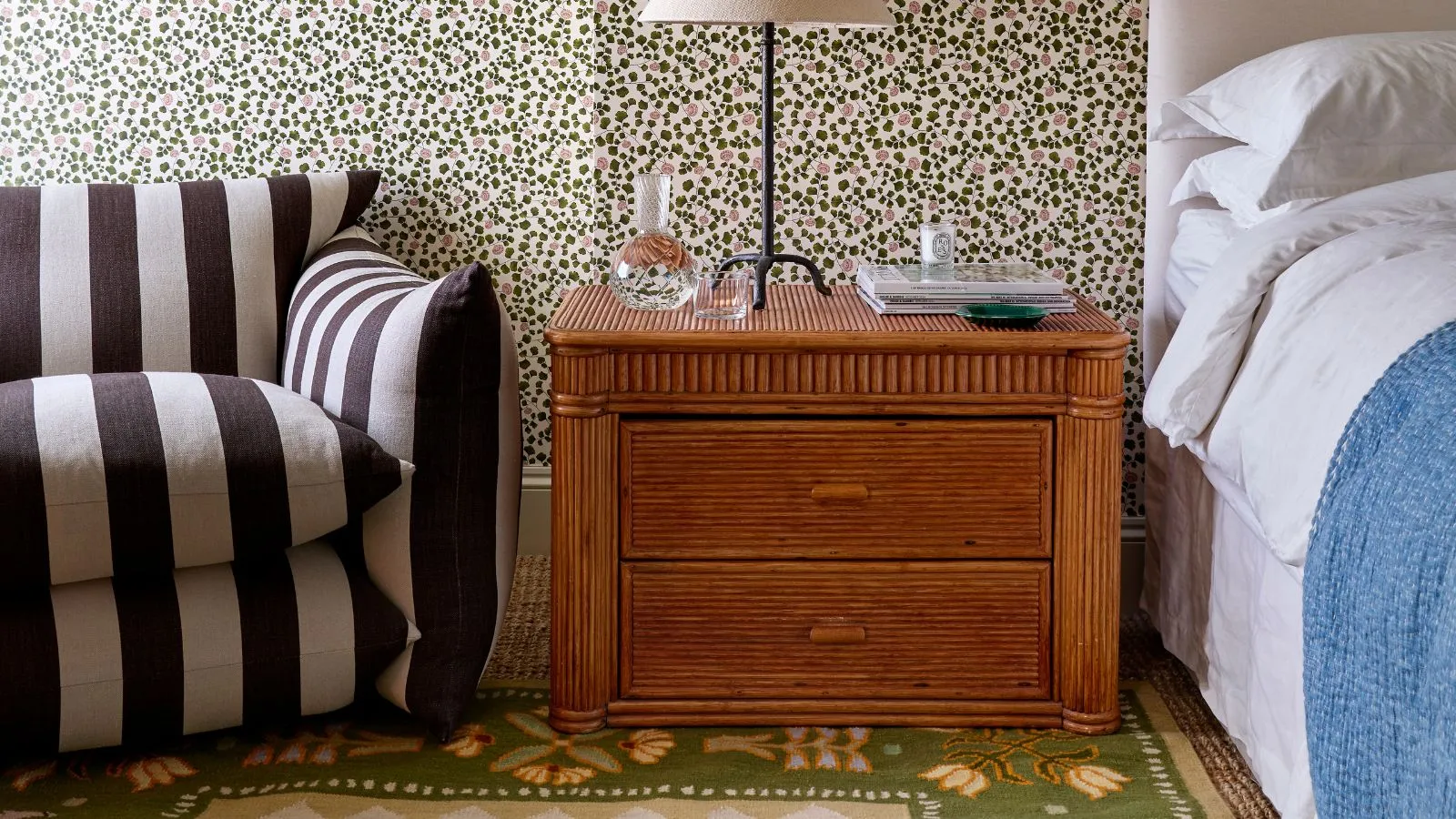 Die Teppichkollaboration von Lucy Williams Home und Pelican House zeigt einen grünen, floralen Flachgewebeteppich in einem Schlafzimmer mit Blumentapete, weißer Bettwäsche, einem Vintage-Nachttisch aus Bambus und einem schwarz-weiß gestreiften Stuhl' loading='lazy' title=