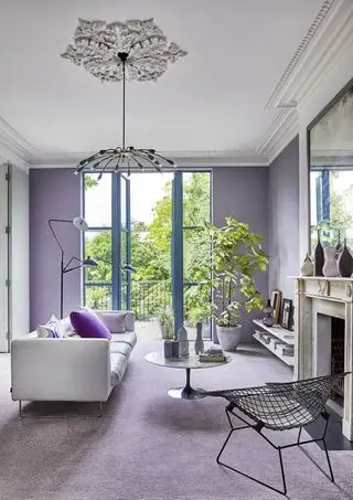 salón con alfombras y paredes lilas, sofá blanco, sillón de metal, plantas, mesa de centro redonda, lámpara de pie