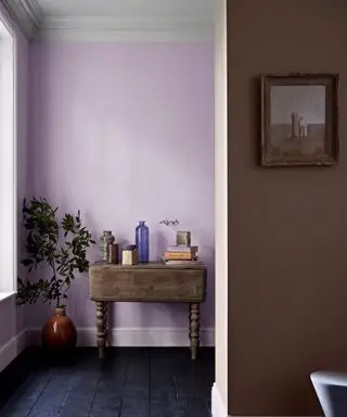 Esquina de la habitación, paredes pintadas de color malva, una pequeña consola de madera decorada con adornos, suelos de madera negros y otras paredes pintadas de marrón