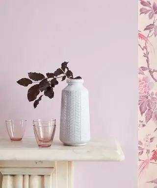 Primer plano de una pared pintada de color malva encima de la chimenea, papel tapiz floral de color púrpura a la derecha, jarrón blanco con flores secas de color púrpura, pequeños vasos rosados