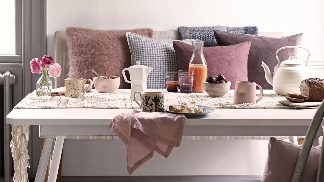 Una escena de desayuno brillantemente iluminada presenta una mesa de comedor blanca cubierta con un camino de macramé color crema y una variedad de cerámicas estampadas y de color rosa suave. Sobre la mesa hay una tetera blanca, una cafetera Moka, una jarra de zumo, tazones de fruta y cereales y un pequeño jarrón de cristal con ranúnculos rosas. Detrás de la mesa, hay un banco decorado con varios cojines de felpa en tonos rosa malva y azul grisáceo. La estética general es aireada y acogedora y se apoya en una paleta de colores suaves y románticos.' loading='lazy' title=
