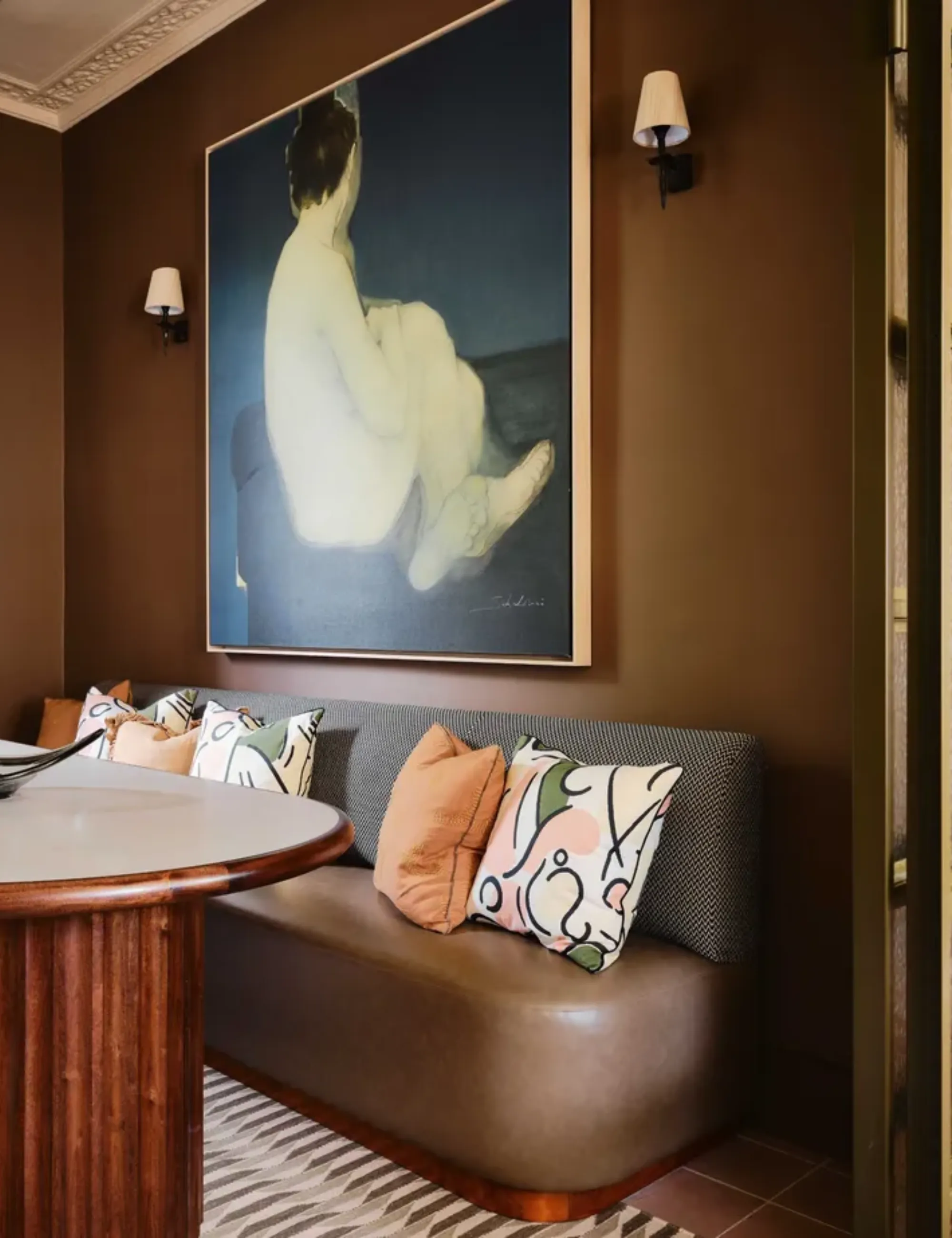 Una sala da pranzo marrone con un tavolo in legno curvato con base scanalata e un accogliente divanetto imbottito e un grande dipinto incorniciato sul muro' loading='lazy' title=