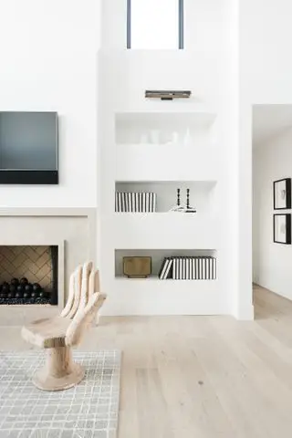 Soggiorno con pavimento in legno chiaro, pareti bianche e scaffali integrati, camino con TV sopra, sedia in legno intagliato a forma di mano e tappeto con motivi neutri