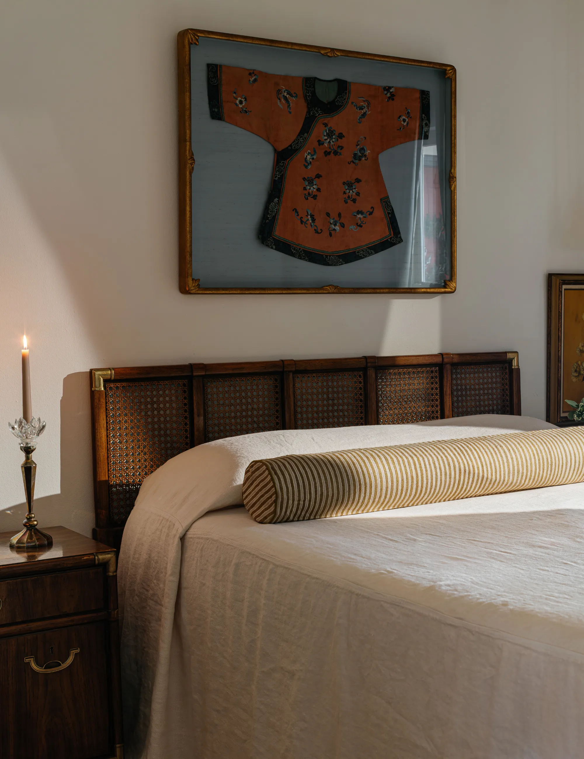 Chambre minimaliste avec art mural kimono, table de nuit antique et literie blanche associée à un traversin extra long à rayures.' loading='lazy' title=