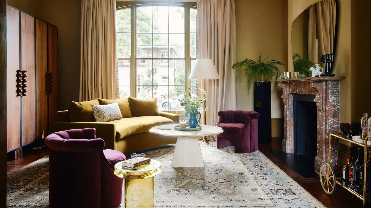 grande sala de estar pintada de ocre com uma antiga lareira de mármore vermelho uma grande janela sofá amarelo curvo mesa de centro redonda branca e poltronas curvas roxas' loading='lazy' title=