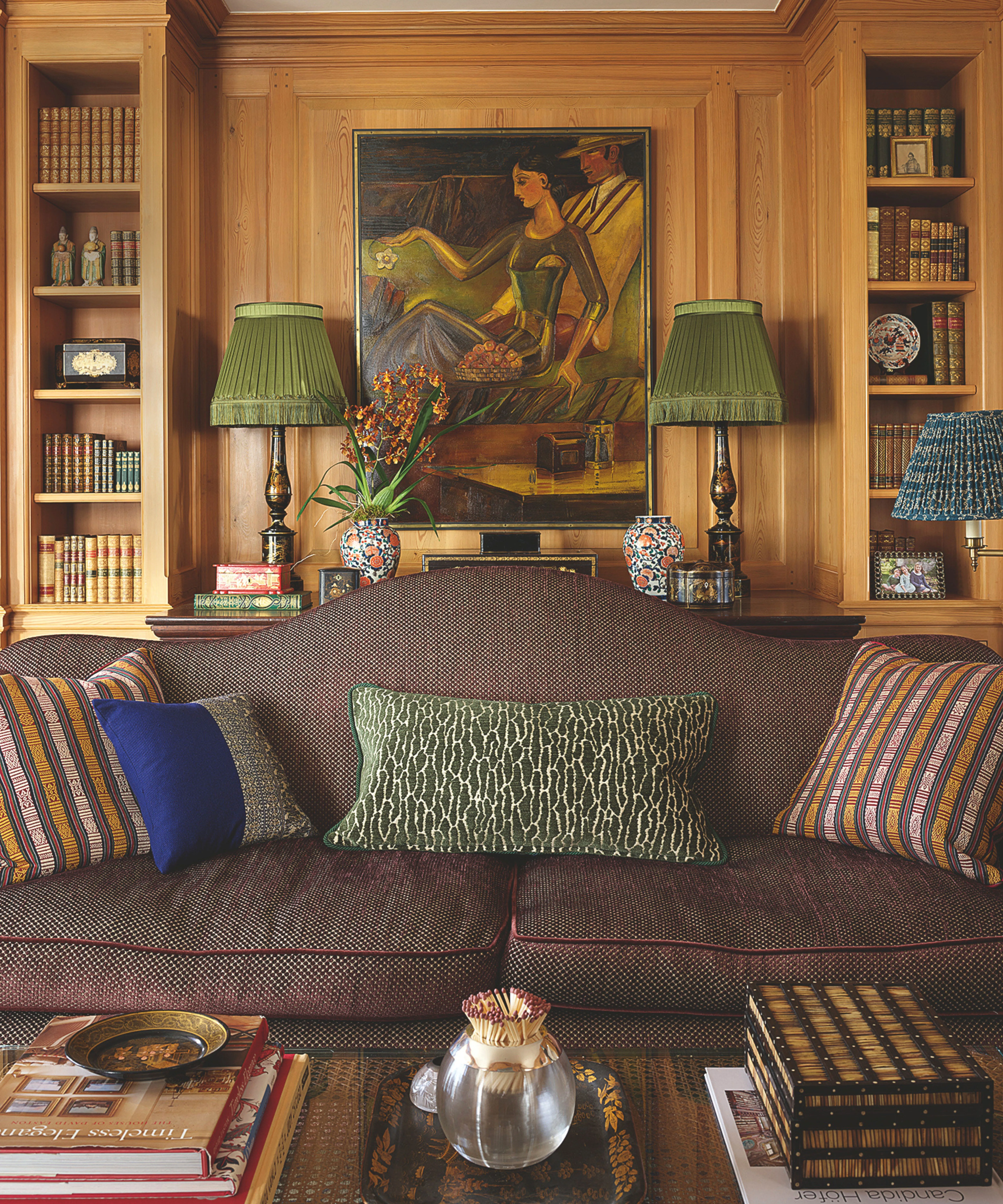 Uma sala luminosa com dois sofás brancos voltados para uma lareira de pedra. Uma grande árvore fica no canto e prateleiras com cerâmica alinham-se na lareira. Um lustre redondo de ouro está pendurado acima e altas janelas com moldura preta permitem a entrada de luz natural. Uma mesa de centro com livros e flores fica no centro.' loading='lazy' title=