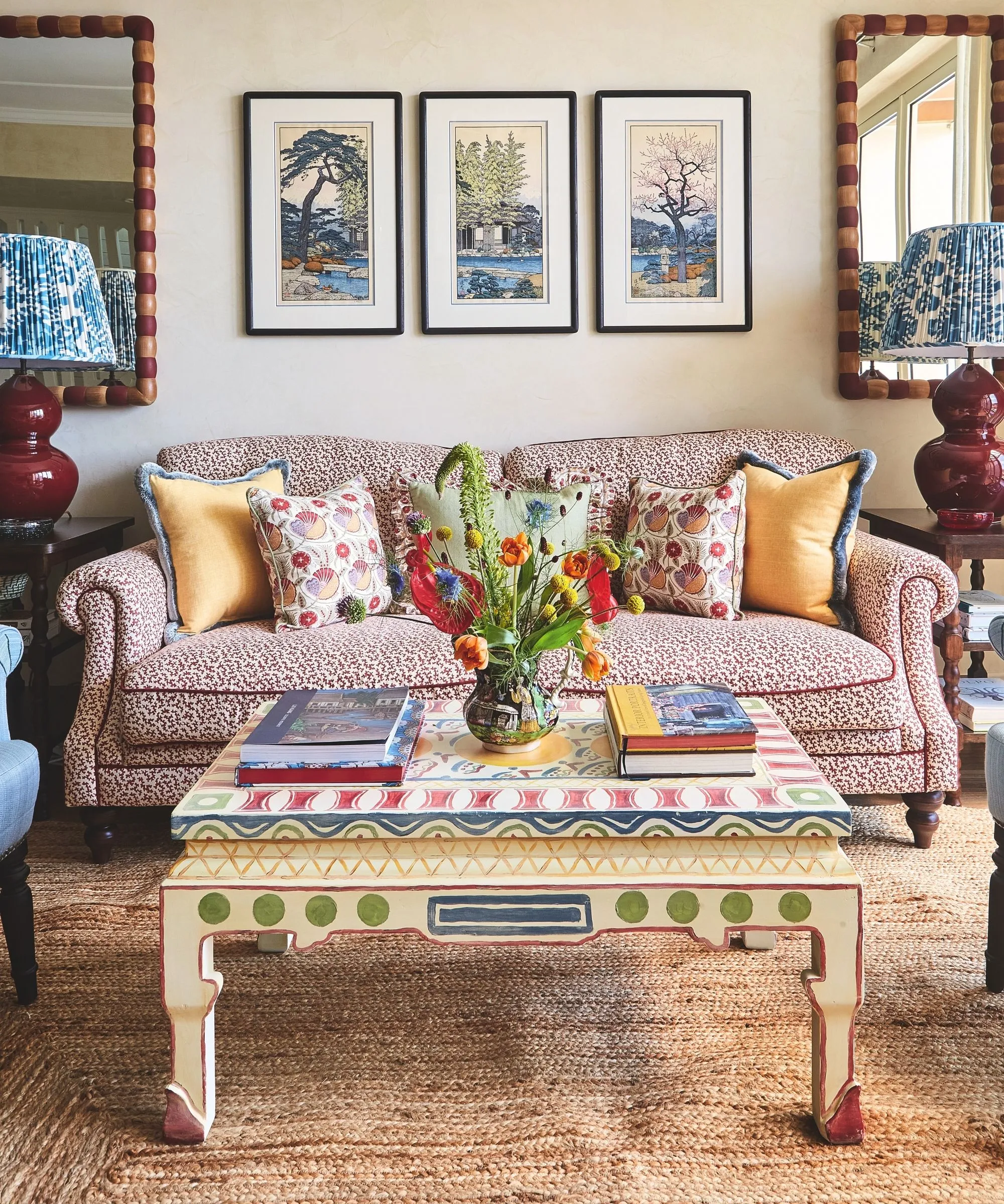 sala de estar com sofá e poltrona estampados e tapete texturizado de ervas marinhas e mesa de centro de madeira pintada em cores e arte nas paredes atrás com espelhos simétricos' loading='lazy' title=
