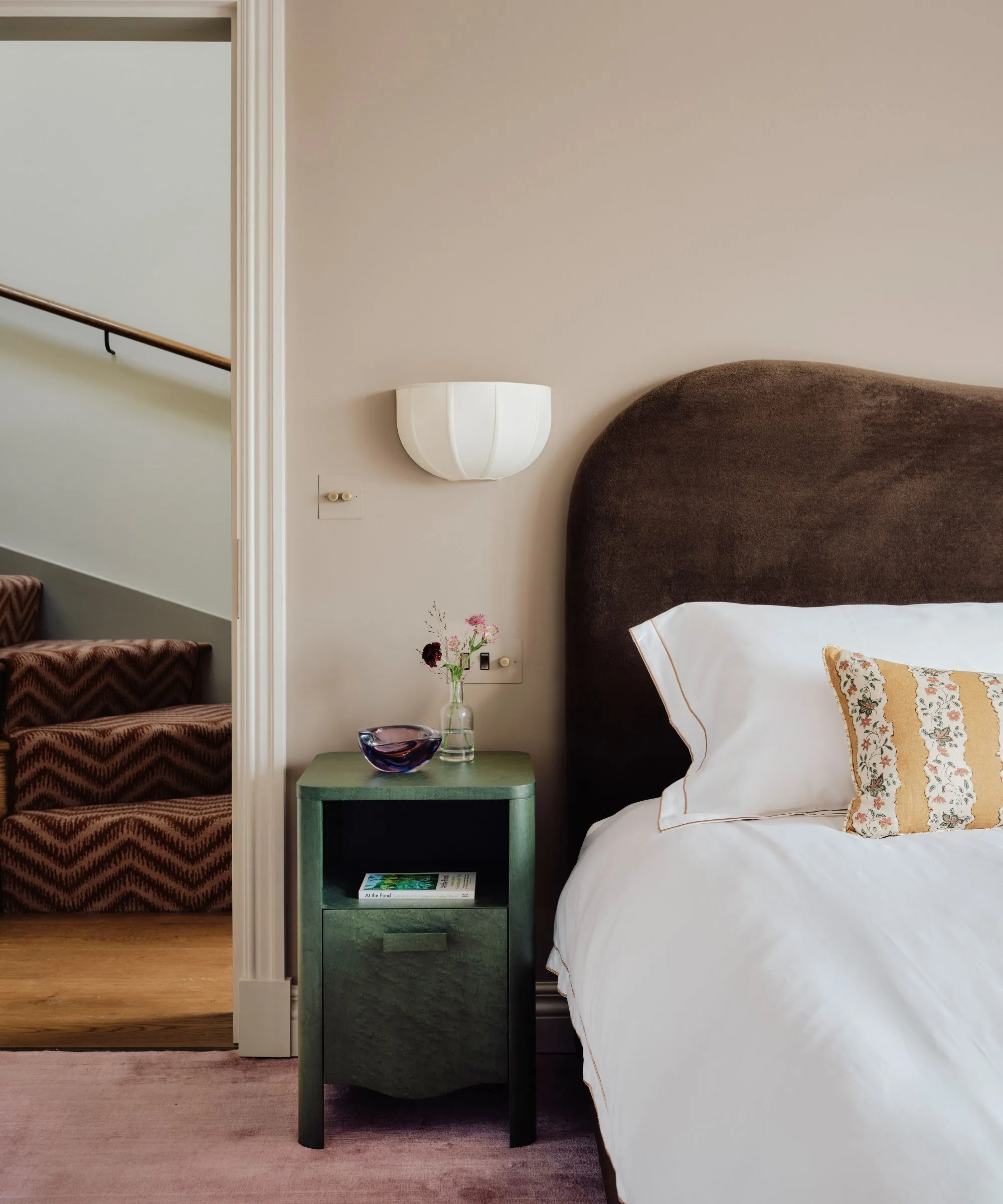 Warmes rosafarbenes Schlafzimmer mit samtigem, schokoladenbraunem Kopfteil, weißer Bettwäsche, gelb gestreiftem Blumenkissen, grünem Nachttisch und lila Teppich' loading='lazy' title=