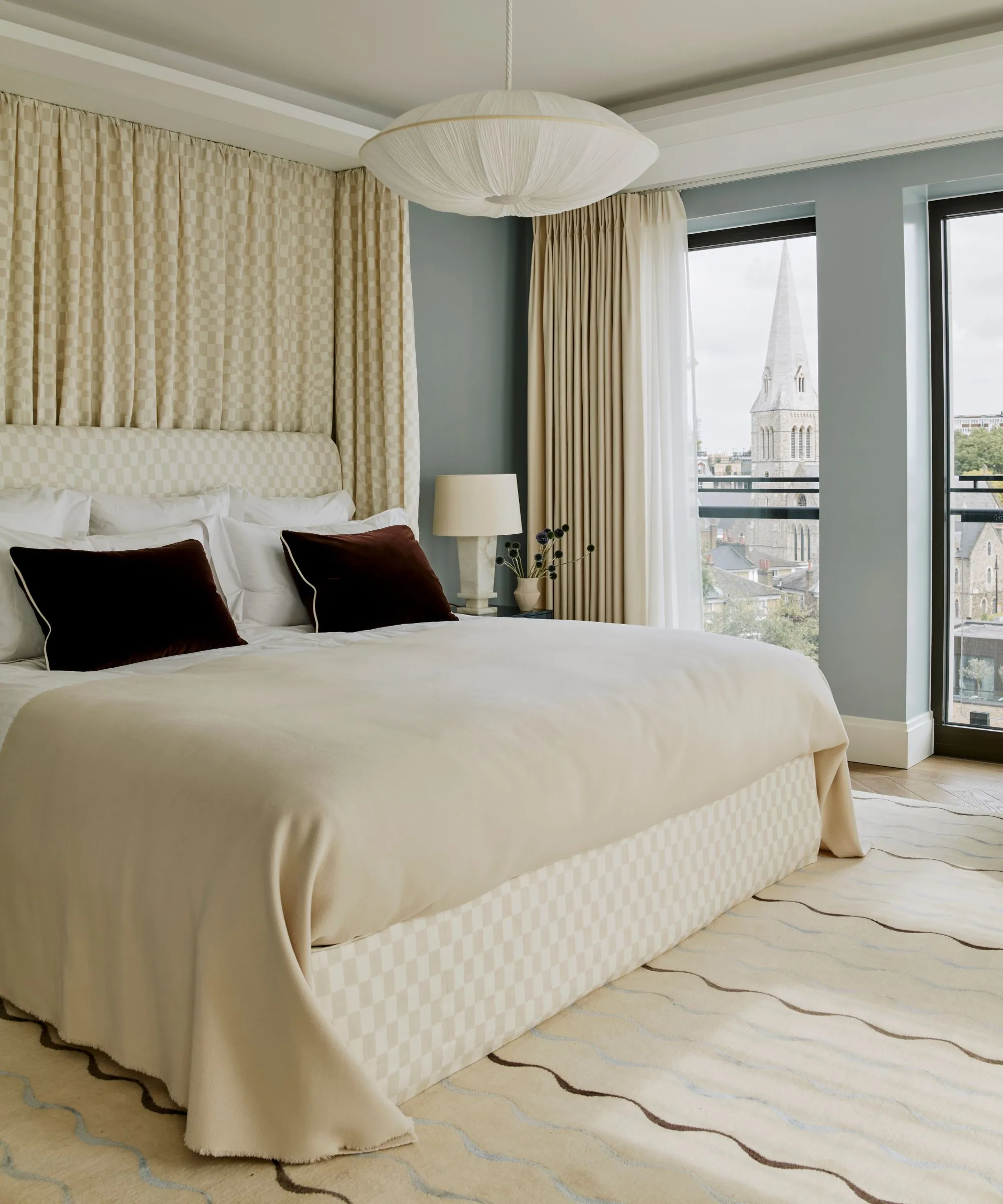 Gemütliches, modernes, hellblaues Schlafzimmer mit großen Glasfenstern, einem Himmelbett-Teppich mit Wackeldruck und burgunderroten Kissen auf einer neutralen Bettwäsche und einem Überwurf' loading='lazy' title=