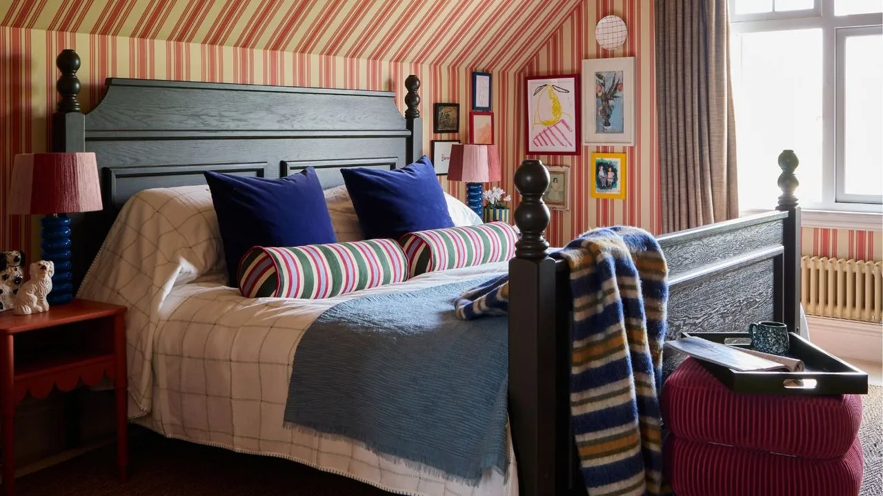 Schlafzimmer mit schräger Decke und beige-rot gestreifter Tapete, die bis über die Decke reicht. In der Mitte steht ein großes dunkles Vintage-Holzbett mit gestreiften Überwürfen und Kissen sowie ein Samthocker am Fußende des Bettes' loading='lazy' title=