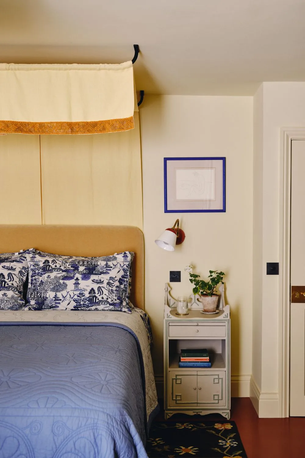 Weiches, buttergelbes Schlafzimmer mit einem gelben Betthimmel, ockerfarbenem Samtkopfteil und blauer Toile-Bettwäsche mit einem Vintage-Nachttisch' loading='lazy' title=