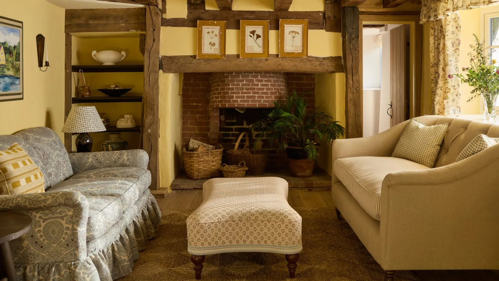 Gemütliches Wohnzimmer im Landhausstil mit buttergelben Wänden, einem gemauerten Kamin und einem blau gemusterten Sofa mit Sockel, einem gelb karierten Kissen und einem gemusterten Ottoman' loading='lazy' title=