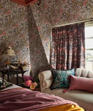 Geblümtes, maximalistisches, auffälliges Schlafzimmer mit gemusterten Vorhängen, Tapeten, Polstermöbeln, Bett mit Decken und Kissen, cremefarben gestreiftes Sofa mit Kissen am Fenster