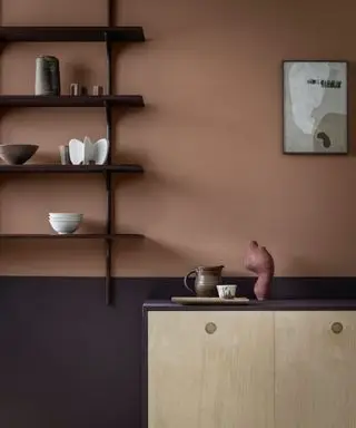 Eine zweifarbige Wand mit hellbrauner Farbe oben und dunkelbrauner Farbe unten, ein offenes Regal aus dunklem Holz mit kleinem Dekor und ein Beistelltisch aus hellem Holz.
