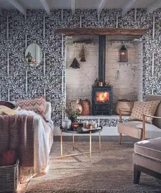 Gemütliches Wohnzimmer oder gemütliches Wohnzimmer mit brennendem Holzofen in einer weiß gestrichenen Nische, Sesseln und Sofa mit Kissen und Stoffüberwürfen. Natürliche, herbstliche Farben und Töne.
