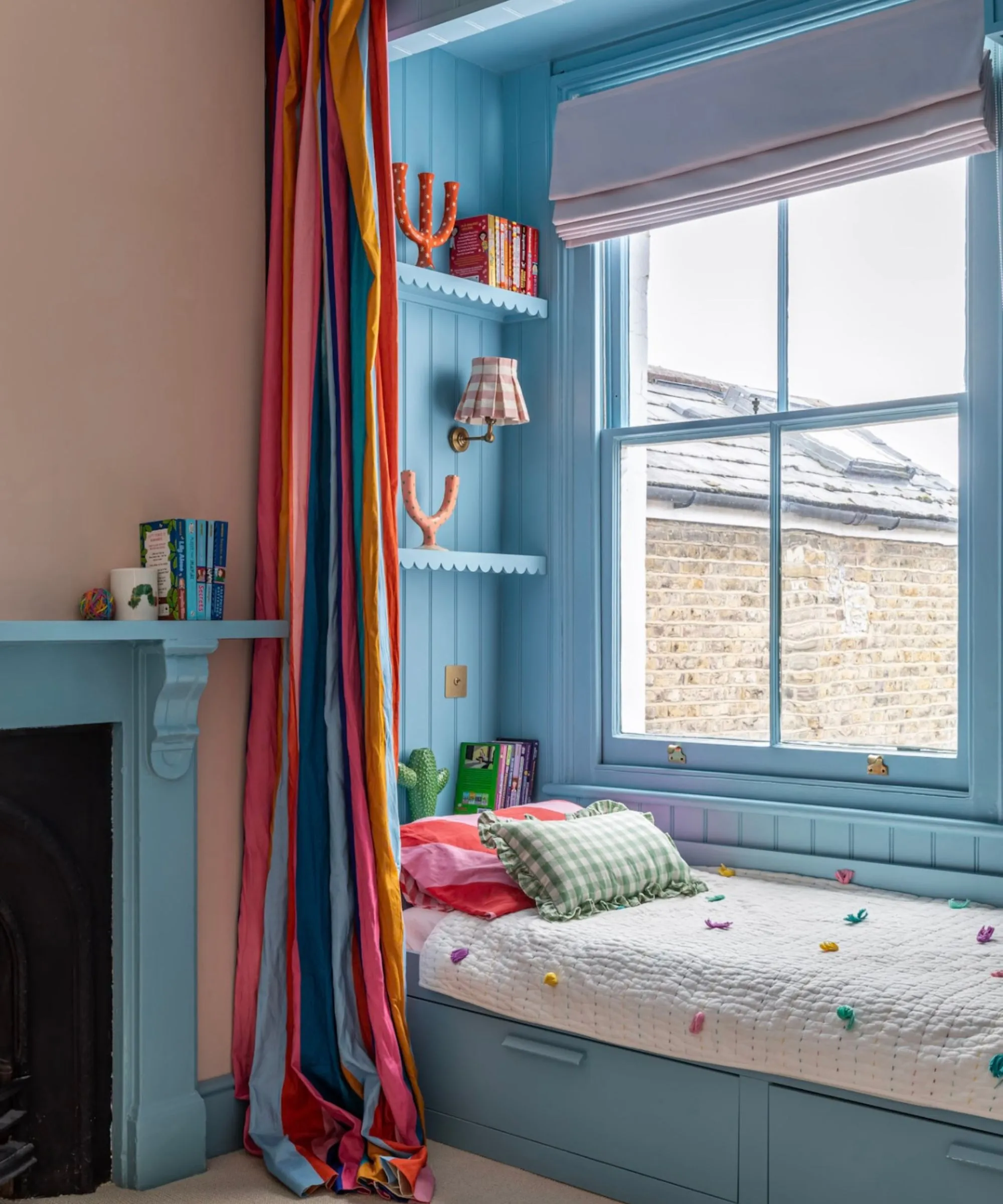 Dětský pokoj se sádrově růžovými stěnami, modrá kabinová postel vedle okna s barevným závěsem a modrým krbem.' loading='lazy' title=