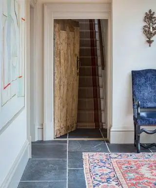 Spazio interno tradizionale con pavimento in pietra ardesia, pareti dipinte color crema con opere d'arte e tradizionali candelabri in metallo, che si affaccia su una porta rustica in legno aperta con scala rivestita in moquette, sedia occasionale in velluto blu e tappeto a motivi multicolori