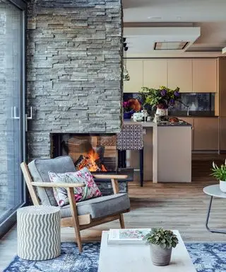 Zona giorno a pianta aperta con parete in pietra ardesia e caminetto, pavimento in legno chiaro con tappeto a motivi blu, poltrona lounge in legno con cuscini grigi imbottiti, tavolino rotondo con motivo geometrico, affacciato sulla cucina con isola e armadietti